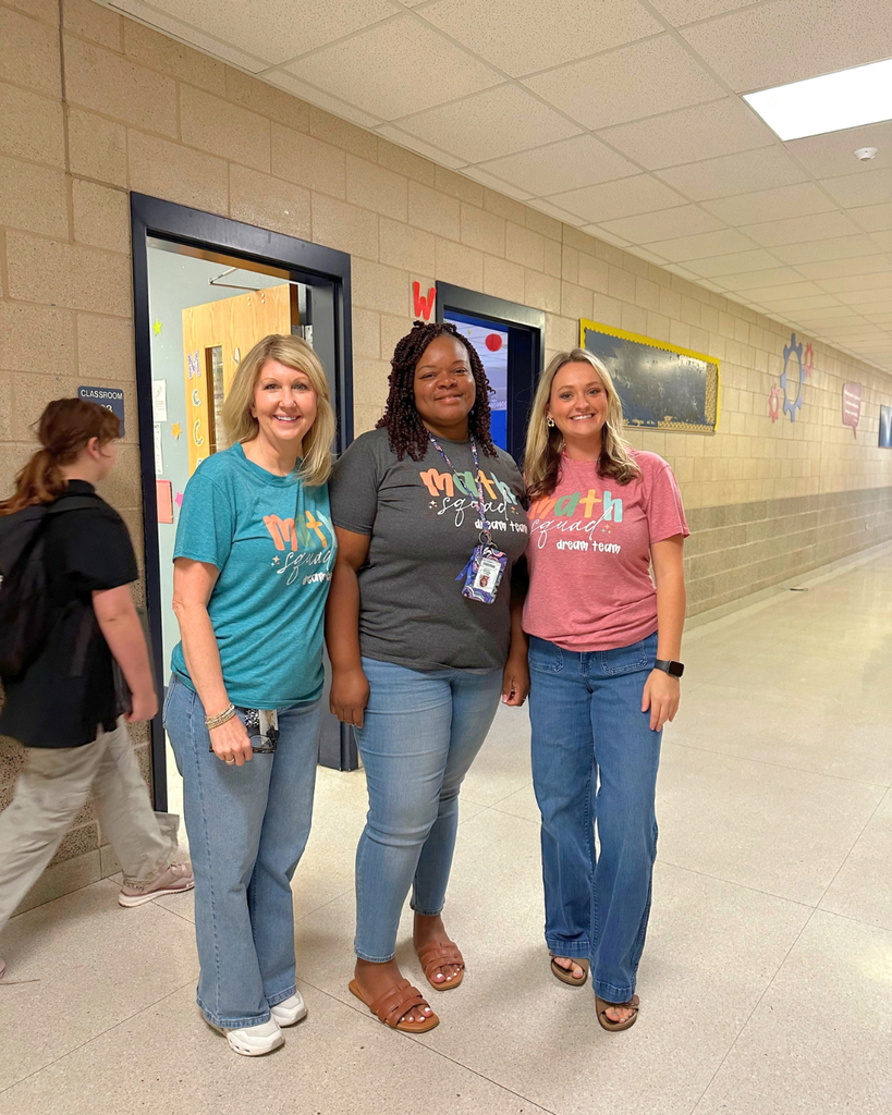 Three smiling women standing together in a school hallway, wearing colorful t-shirts and jeans.