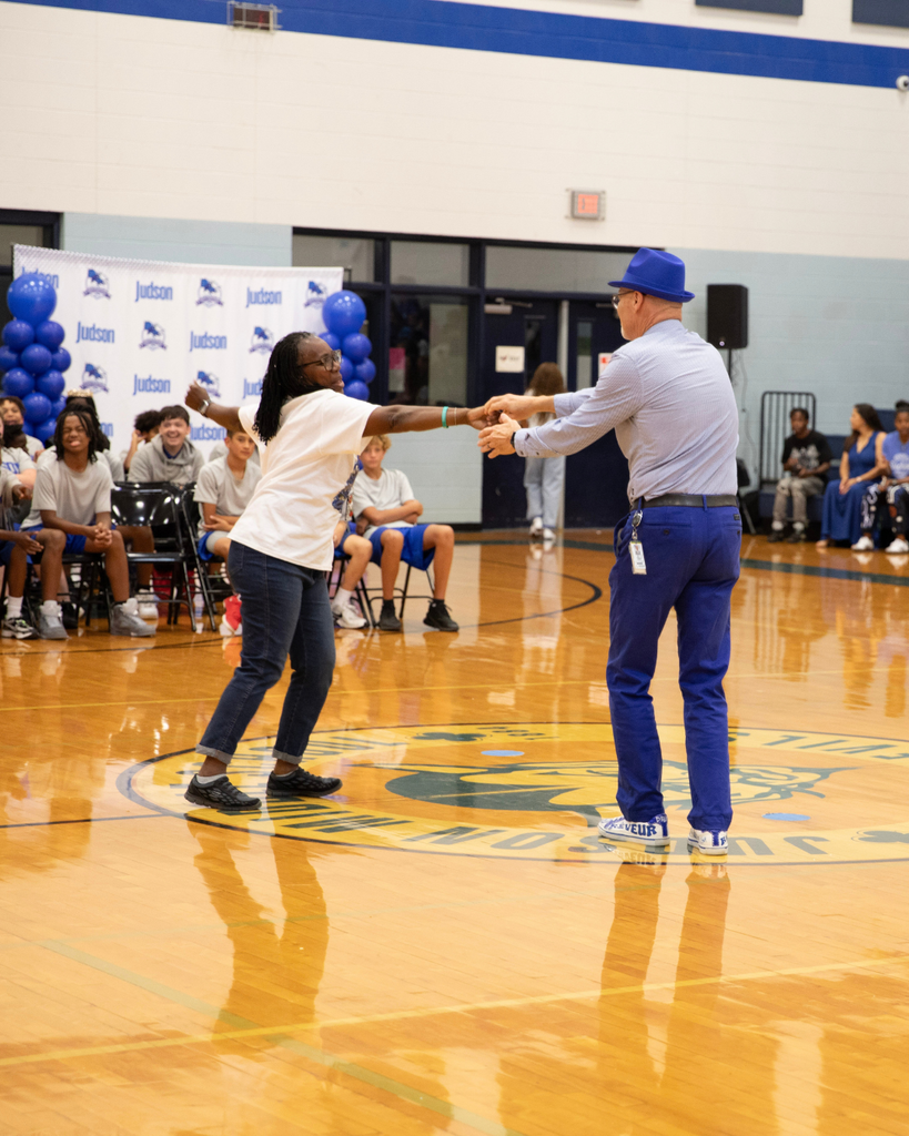 A woman and a man dancing together on a gymnasium basketball court during a school event, with students seated in the background.