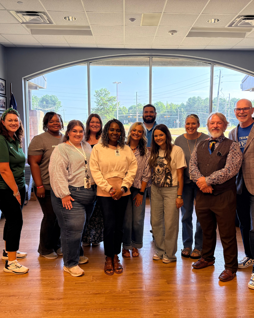 A large group of diverse school staff members smiling and standing together indoors in front of a wide window.