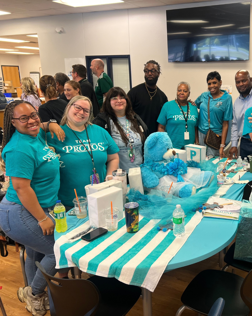 A group of school staff members wearing matching turquoise shirts posing around a decorated table with school spirit items and a mascot head.