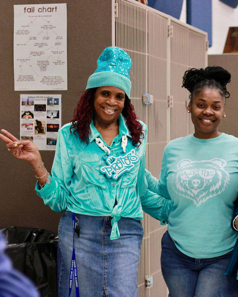 Two smiling women wearing teal school spirit shirts and denim jeans posing together indoors, with one woman flashing a peace sign while wearing a knit blue beanie.