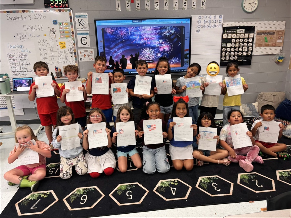A class of first grade students are holding up papers that have the American flag and sentences they wrote about the flag.