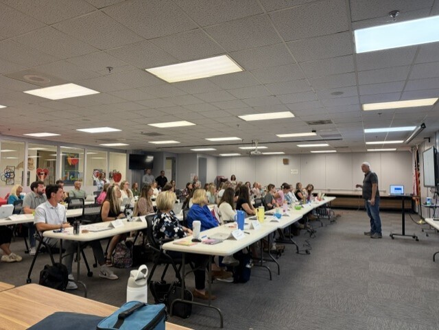 A classroom with rows of desks, attendees seated, and a person standing at the front. Lights on the ceiling.