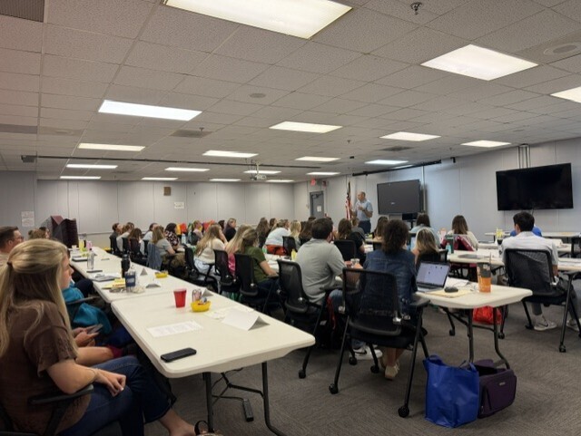 A room with tables, chairs, and a person standing at the front. Some people are seated at tables with laptops, and others are seated on chairs.