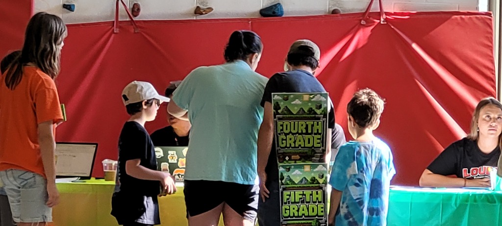 People gather around tables with signs that say "Fourth Grade" and "Fifth Grade" in front of a red wall.