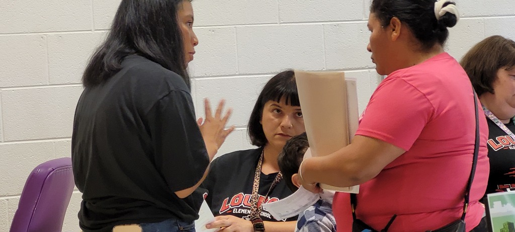 Two women discuss with each other; one holds a piece of paper. Two others sit behind them.