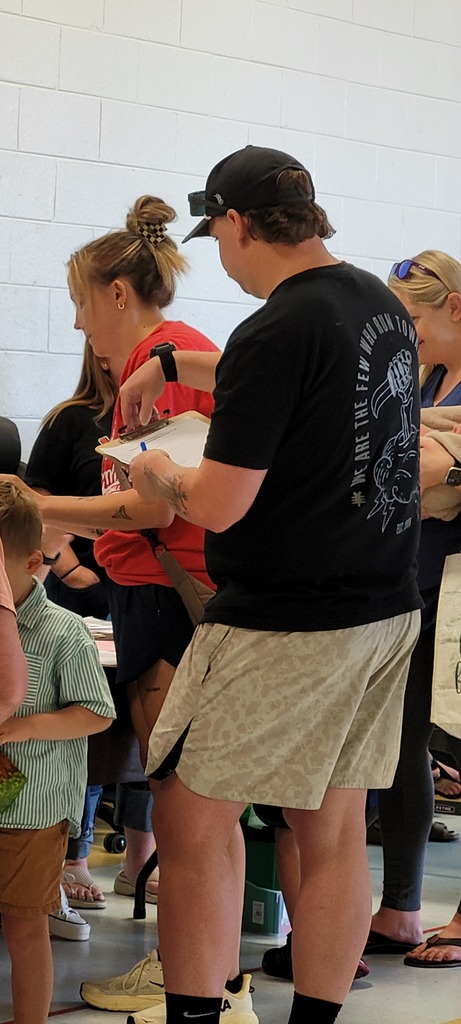 A man in a black shirt and shorts hands a paper to a woman in a red shirt. Several people stand behind them, one holding a paper bag.