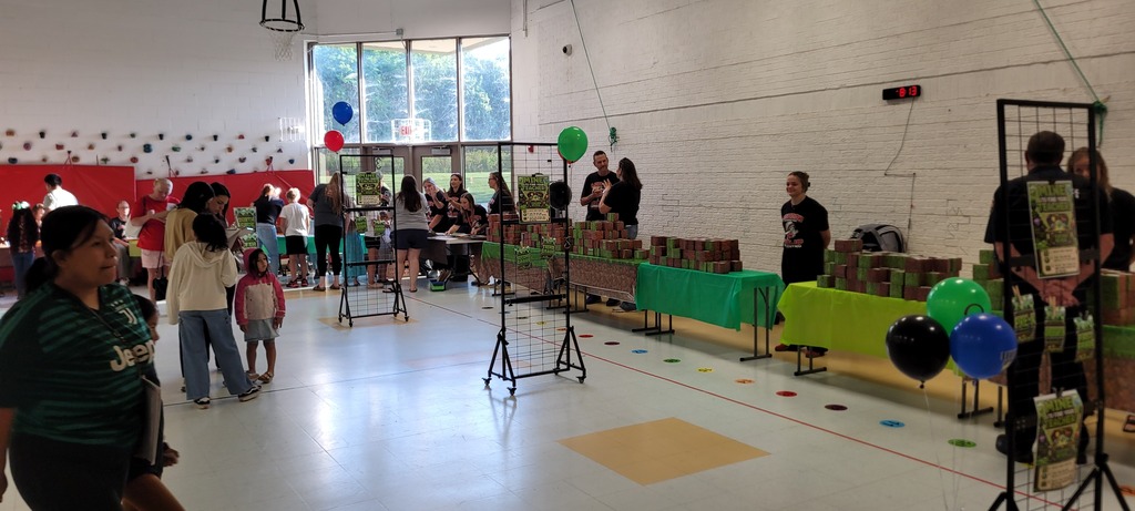 A room with tables and balloons, people walking and standing. Tables are covered with green cloths.