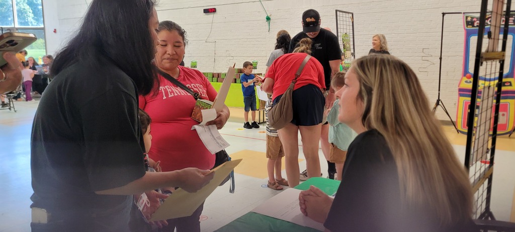 Several adults, some with children, stand in a line to speak with a woman. A few people are standing in the background.