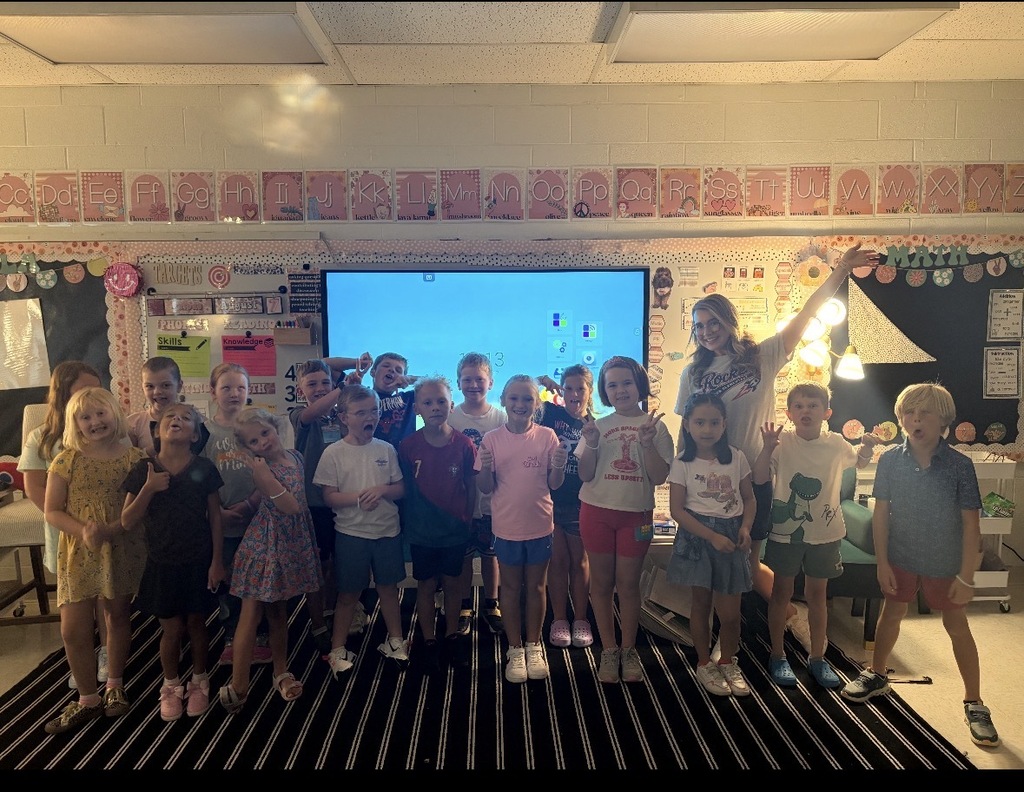 A group of children, with one standing behind a large screen, in a classroom with colorful decorations on the walls.