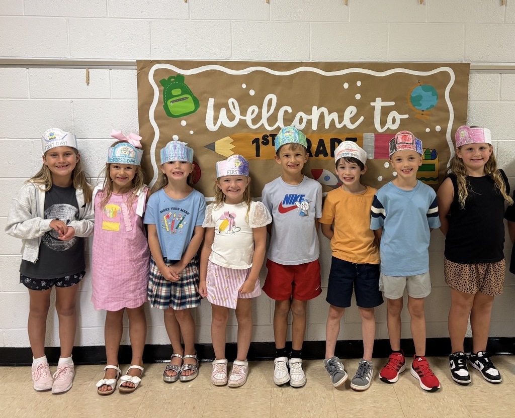 Nine children in hats stand in a row. They are smiling for a picture. A banner behind them says "Welcome to 1st Grad".