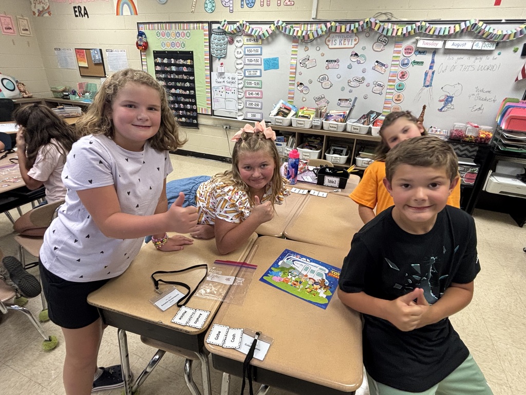 Kids sit at a table in a classroom. A girl in white stands and two kids sit. A board is in the background.
