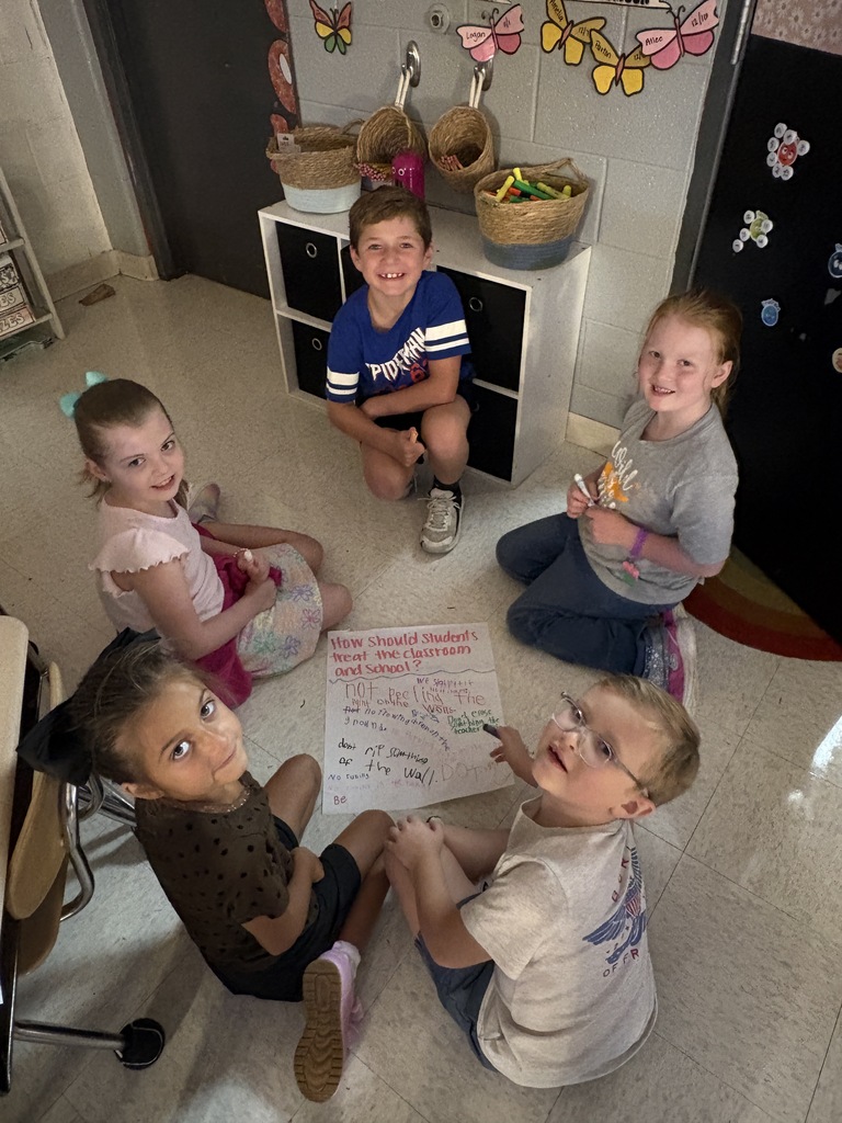 Five children sit in a circle on the floor, with four holding paper. A whiteboard and baskets are in the background.