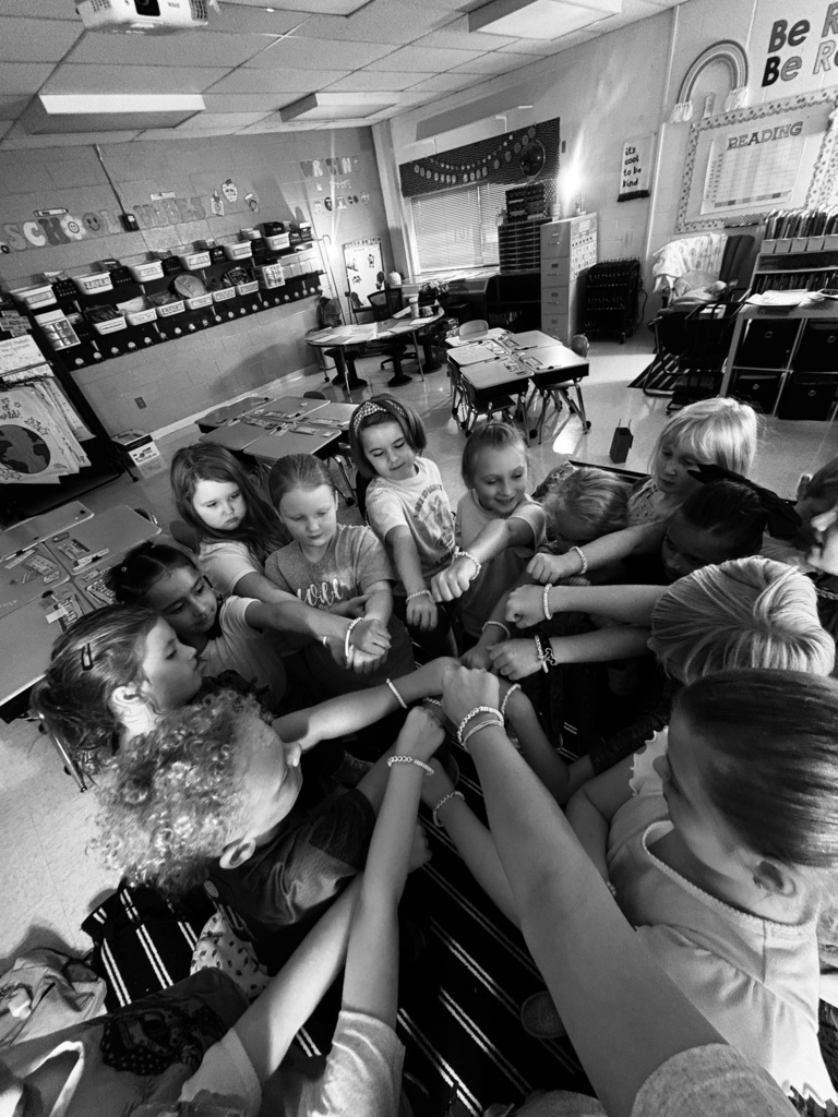 A group of children stand in a circle inside a classroom, holding hands with one another.