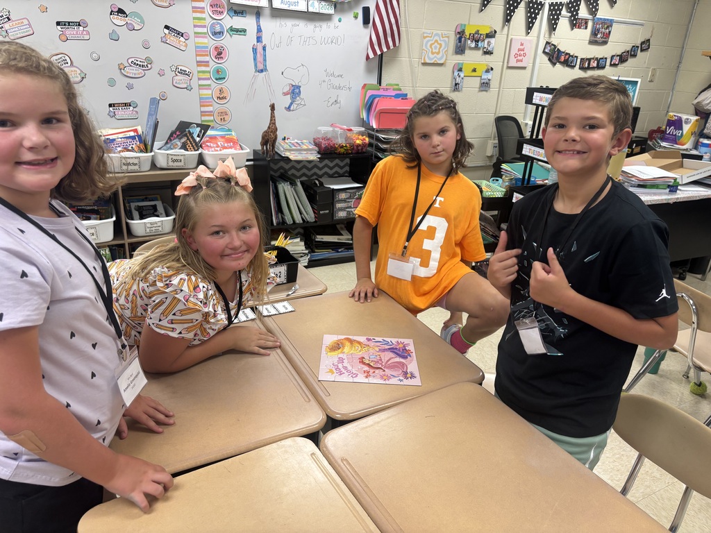 Four children in a classroom, with one leaning on a table, smiling and posing for a photo.