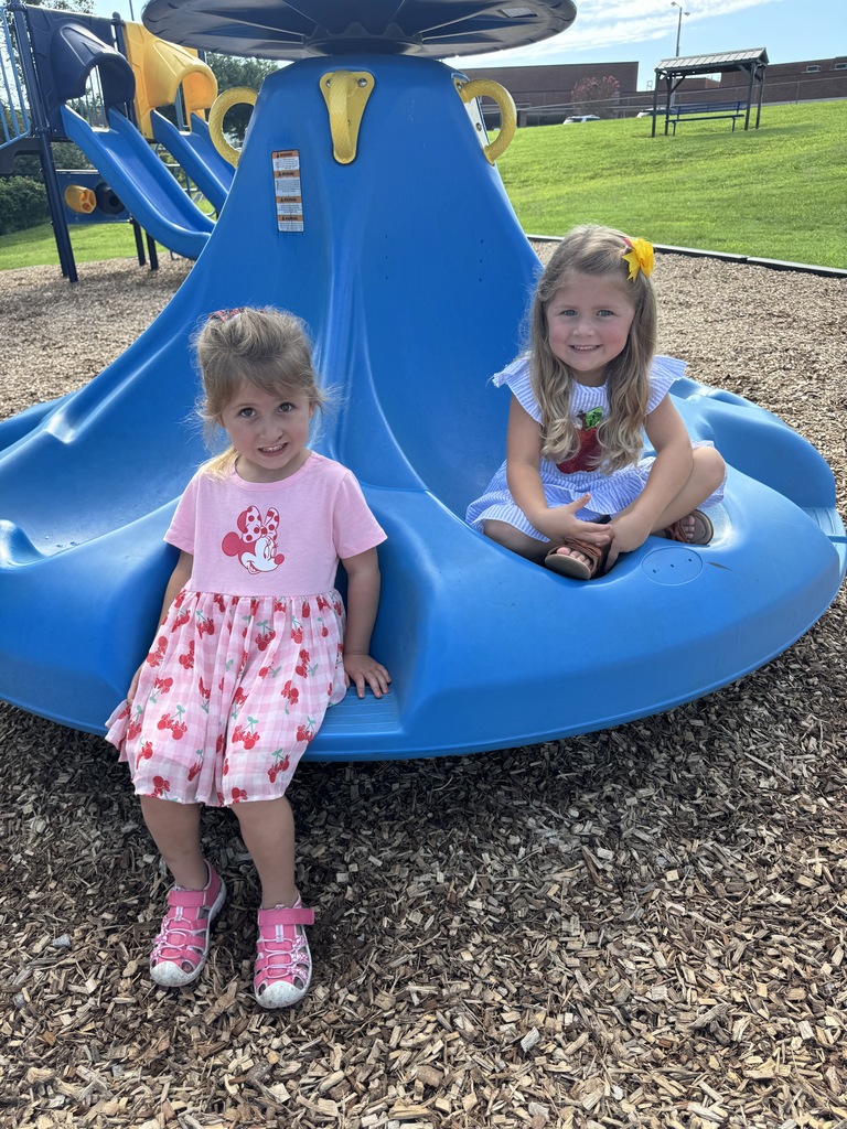 Two girls sit on a blue curved slide; one wears pink shoes and the other yellow hair bows.