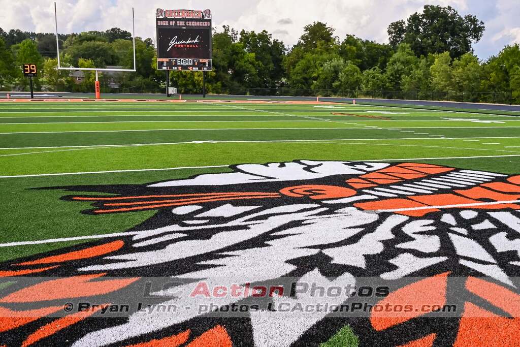 An empty football field with a scoreboard, trees, and a large team logo in the middle.