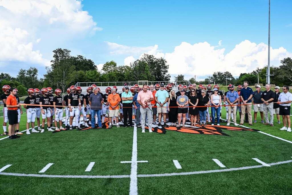 A group of people in sports attire stand on a football field, forming a line. Trees and a cloudy sky in the background.