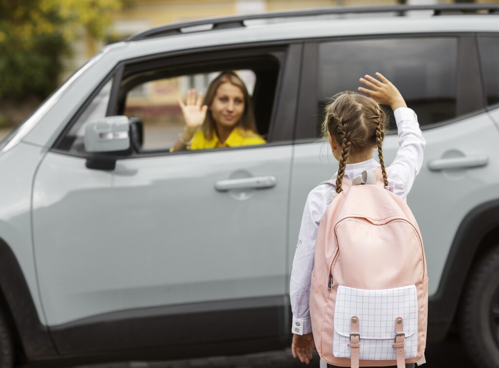 A girl with pigtails waves to a woman in a car, wearing a backpack and standing on the roadside.