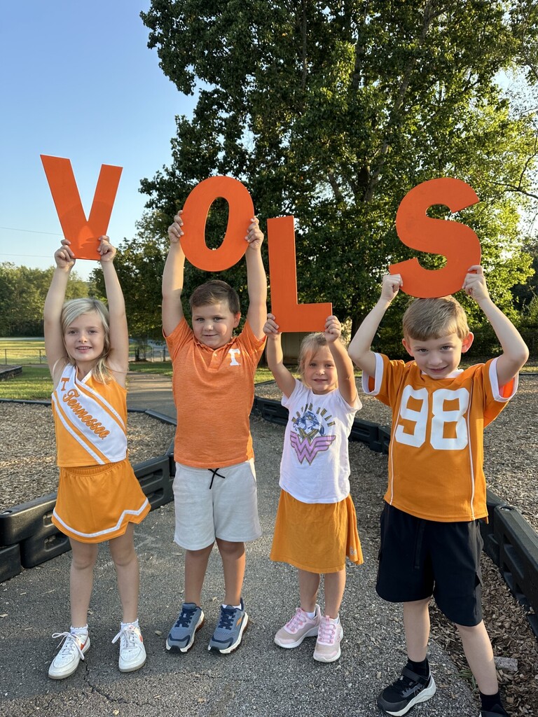 Four children in sports uniforms stand together, holding up letters that spell "VOLS". Behind them, a tree is visible.