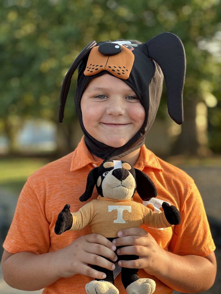 A boy wears a dog hat and holds a stuffed dog in his hands. Trees are in the background.
