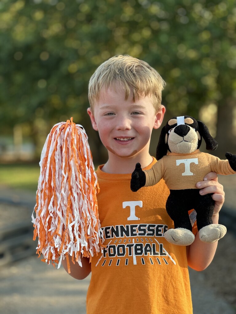 A young boy in an orange Tennessee Football shirt holds a pom-pom and a dog plush toy.
