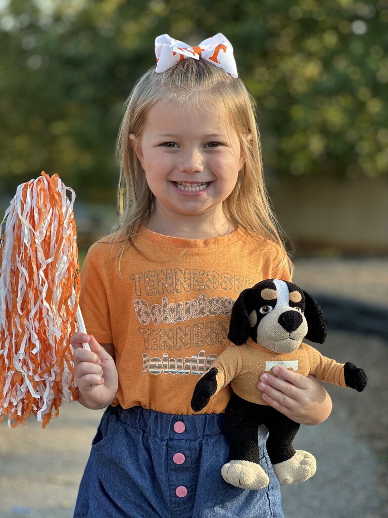 Young girl with blond hair holds a stuffed dog and a pom-pom. She wears orange shirt and blue skirt.