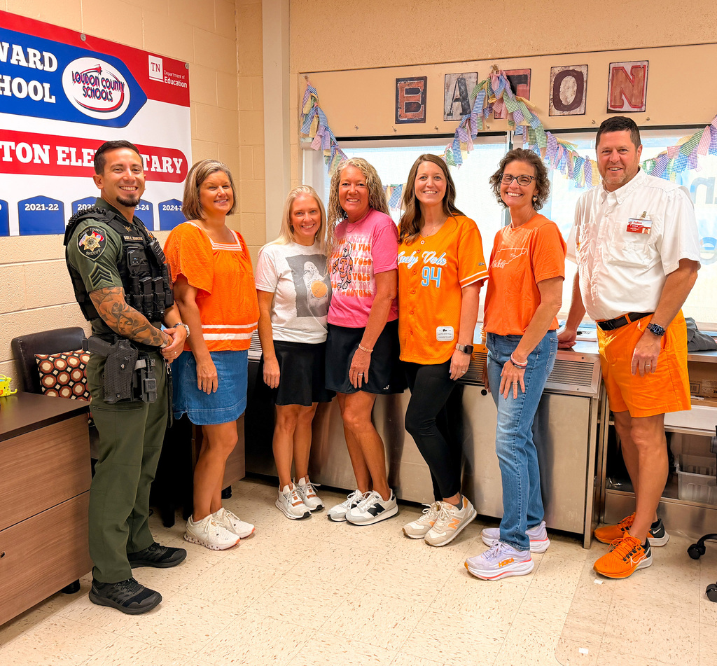 A group of people in orange shirts standing in a classroom. A banner with text is on the wall.