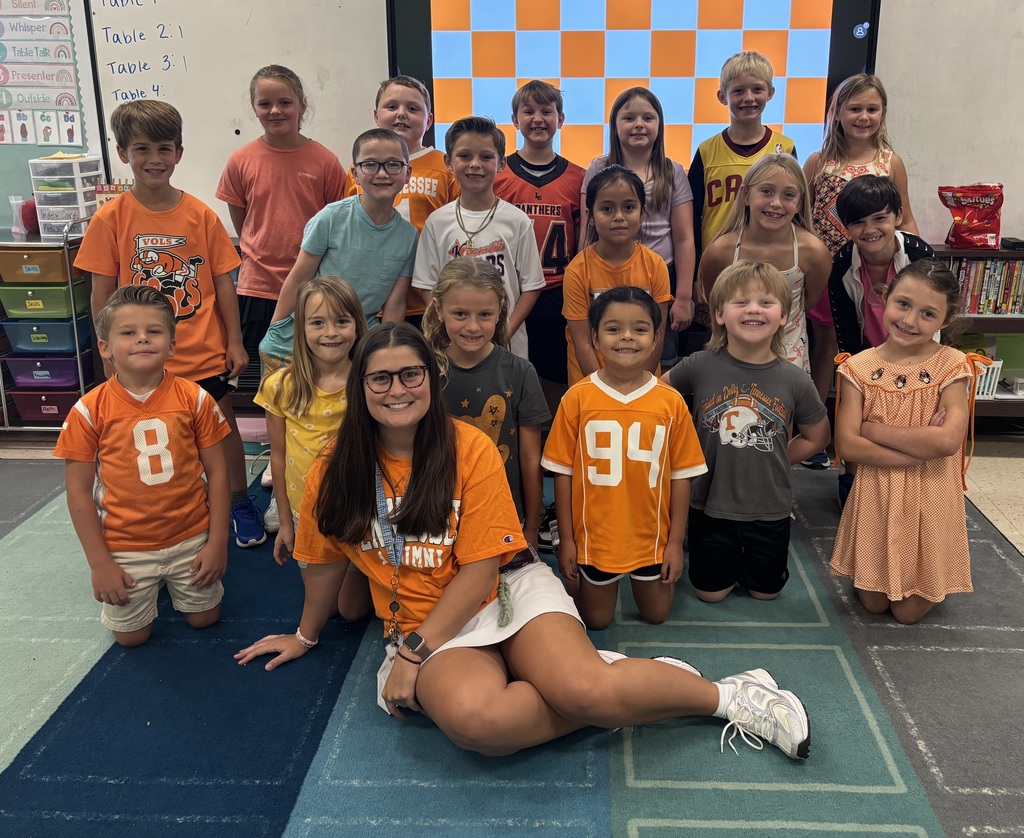 A classroom with students and a teacher; students in orange and gray shirts, one wearing glasses; carpeted floor.