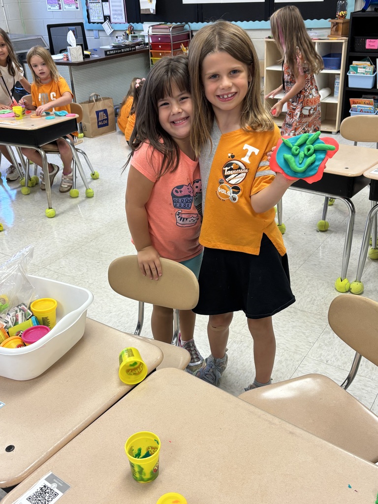 Two girls in a classroom, one wearing a yellow shirt, standing next to a desk filled with cups and other items.