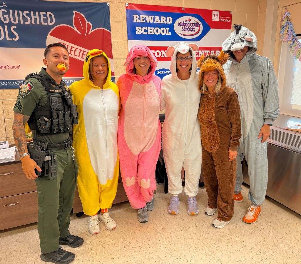 Five people in animal costumes stand together in a classroom, one is a police officer in uniform.