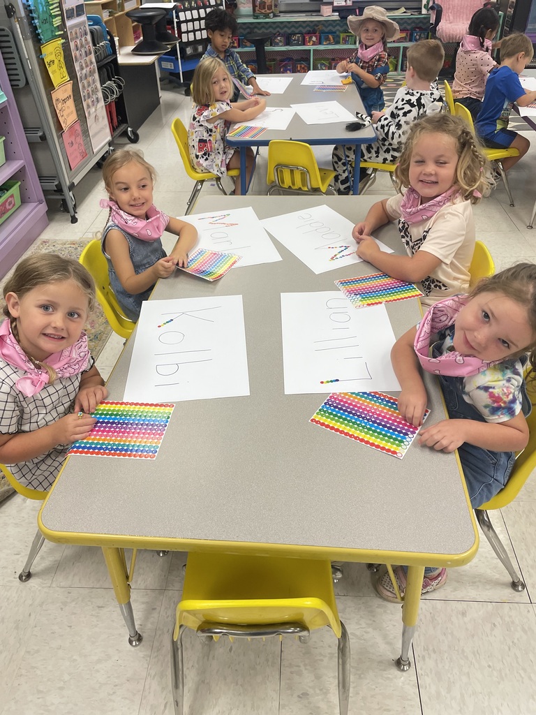 Five young children, wearing bandanas, are sitting around a table, working on papers.