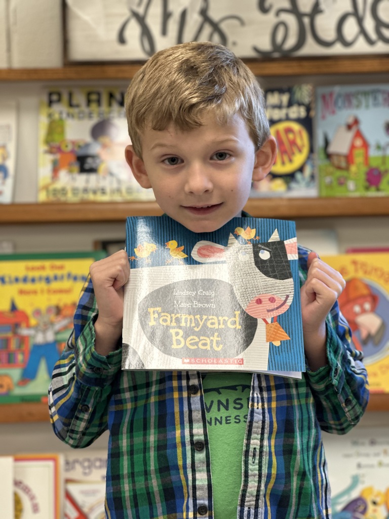 A young boy holds a book titled "Farmyard Beat" in a room with shelves full of colorful books.