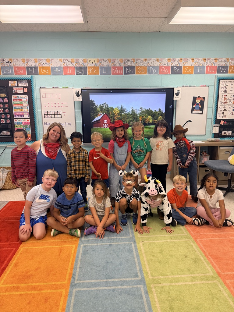 Group of children in a classroom. One child is dressed as a cow. Multi-colored floor with rugs.
