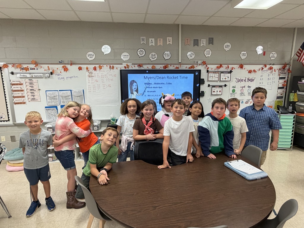 A group of children in a classroom standing around a table, a whiteboard in the background.