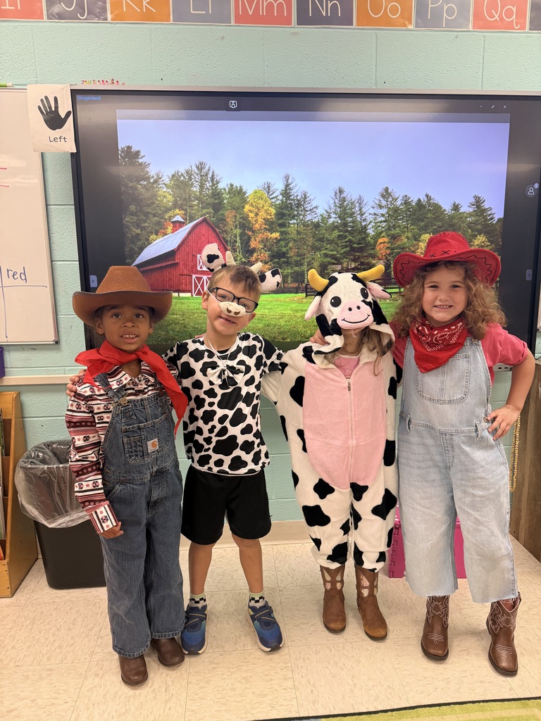 Four kids in costumes, including cowboy hats, stand in a classroom. One wears a cow outfit. A large screen behind them shows a farm.