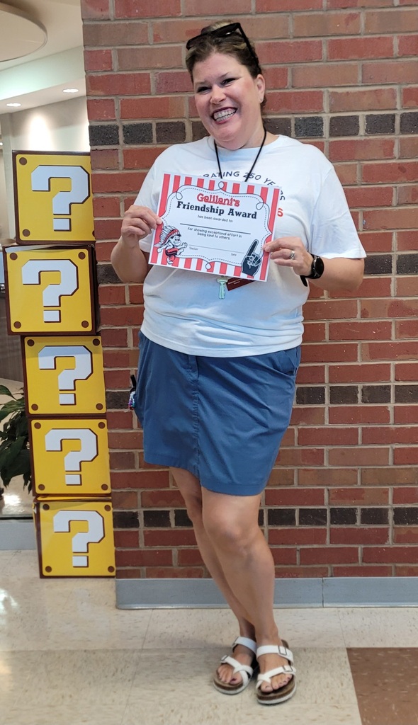 A smiling woman in a white shirt and blue skirt holds a red certificate in front of a brick wall.