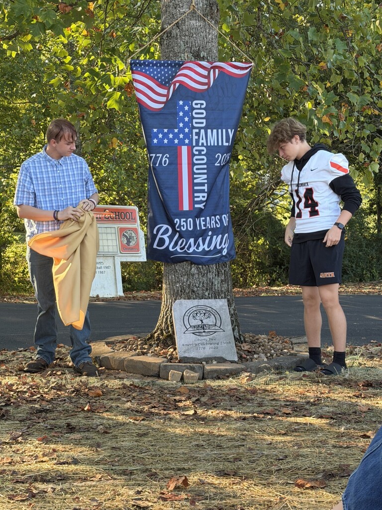 Two young men near a tree, one holding a flag, with a banner reading "GOD COUNTRY FAMILY 250 YEARS OF BLESSING".