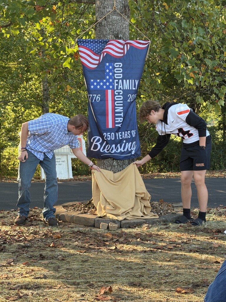 Two people stand in front of a tree with a banner that reads "250 Years of Blessing." They hold a cloth.