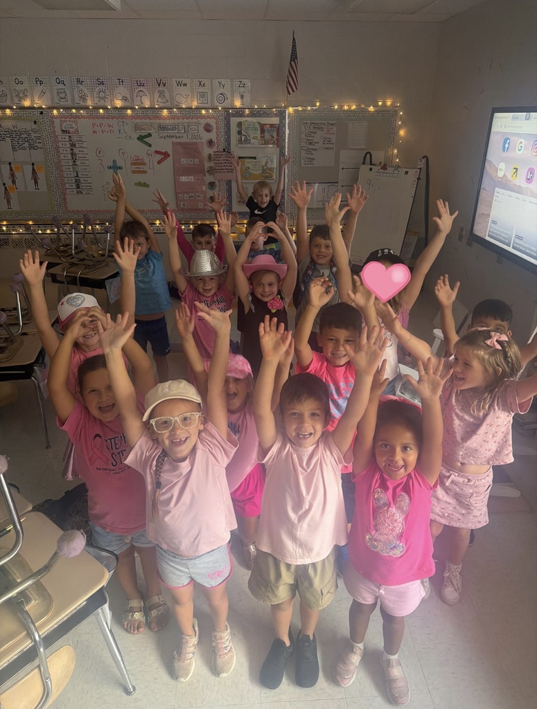 Young children in pink shirts and shorts pose with raised hands in a classroom.