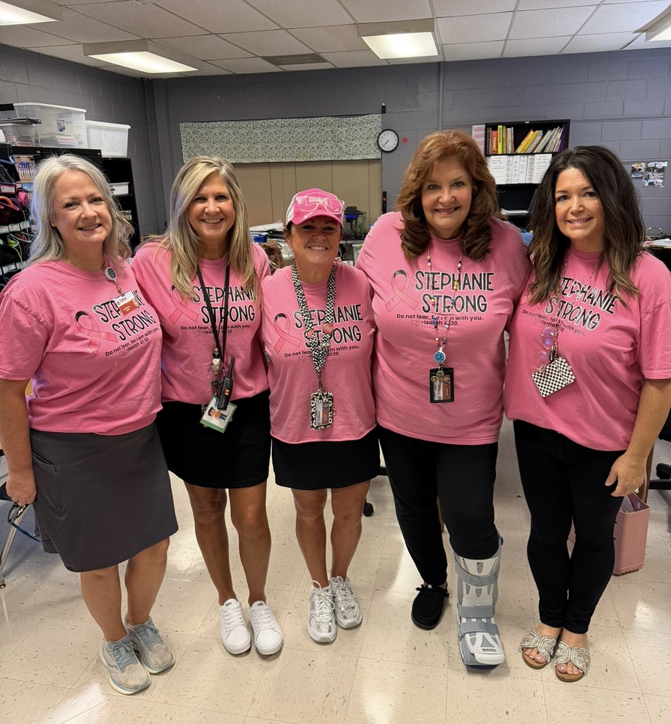 Five women in pink T-shirts pose in an indoor space. They wear sneakers and some have bags.
