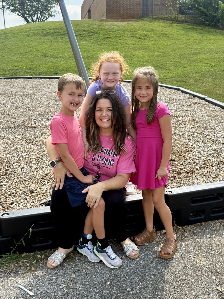 A woman and three children stand on the edge of a playground, smiling and wearing pink shirts.