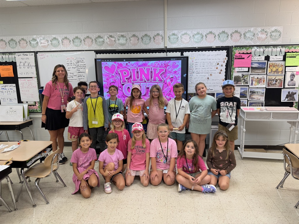A classroom with desks and chairs, children sit and stand, smiling at a camera. A teacher stands behind them. A projector screen shows "PINK OUT."
