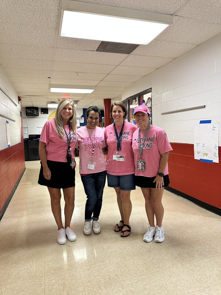 Four women in pink shirts stand in a hallway with red walls and white trim. Each woman wears ID lanyards, and two have pink caps.
