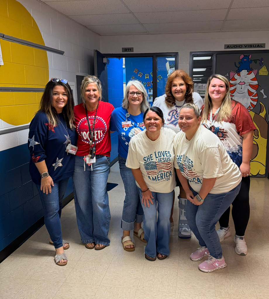 Seven women stand in a hallway, wearing casual clothing, smiling, and posing for a photo.