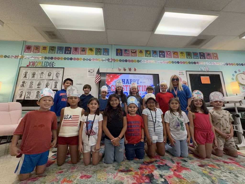 Kids in a classroom pose for a photo wearing chef hats and casual clothing. Bright decorations adorn the walls.