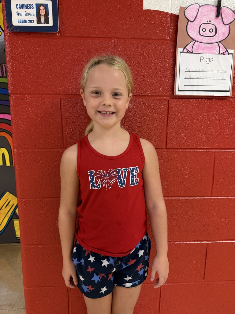 A young girl in a red tank top and shorts stands in front of a red wall with a pig drawing.