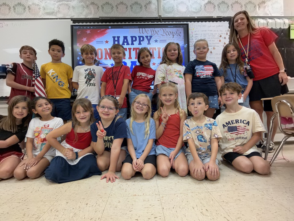 Group of students in a classroom posing for a photo with a teacher, all wearing casual clothes.