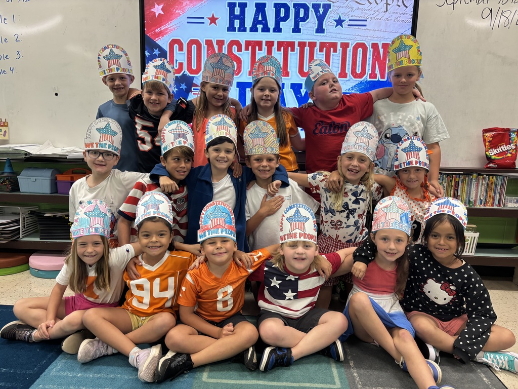 A classroom with students in hats, a white board in the background, and a screen with "Happy Constitution Day".
