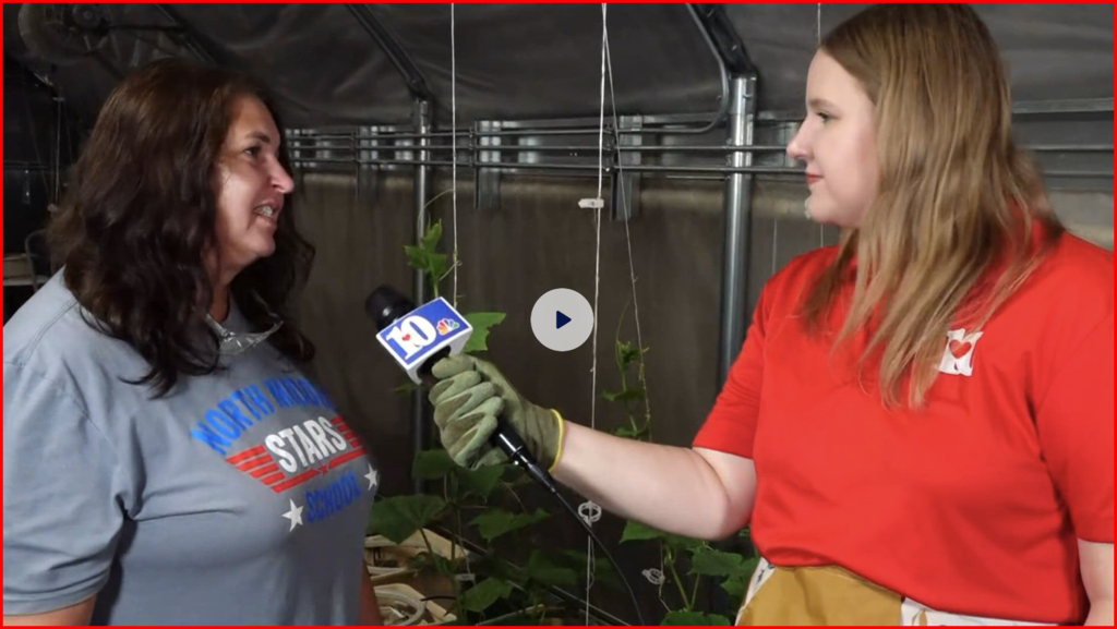 Two women inside a greenhouse, one holding a microphone. The woman on left wears a gray shirt, and the woman on right wears a red shirt.